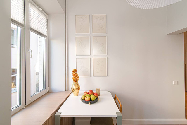 A white dining table stands by the large windows, and framed pictures hang on the wall.