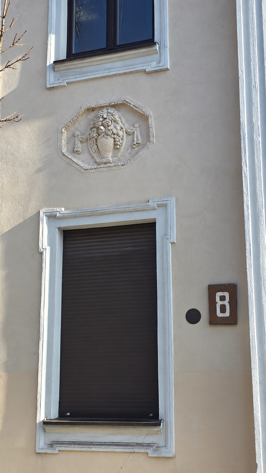 Two windows, a lowered roller blind and a relief depicting a vase with flowers are visible on the building's façade.