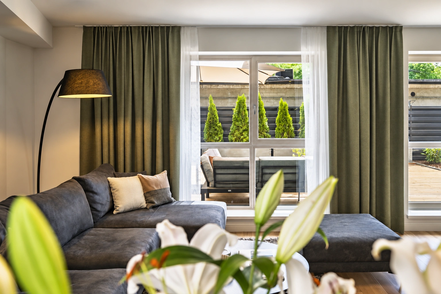 Im Wohnzimmer stehen ein graues Ecksofa und eine Stehlampe, und an den Fenstern hängen lange Vorhänge.