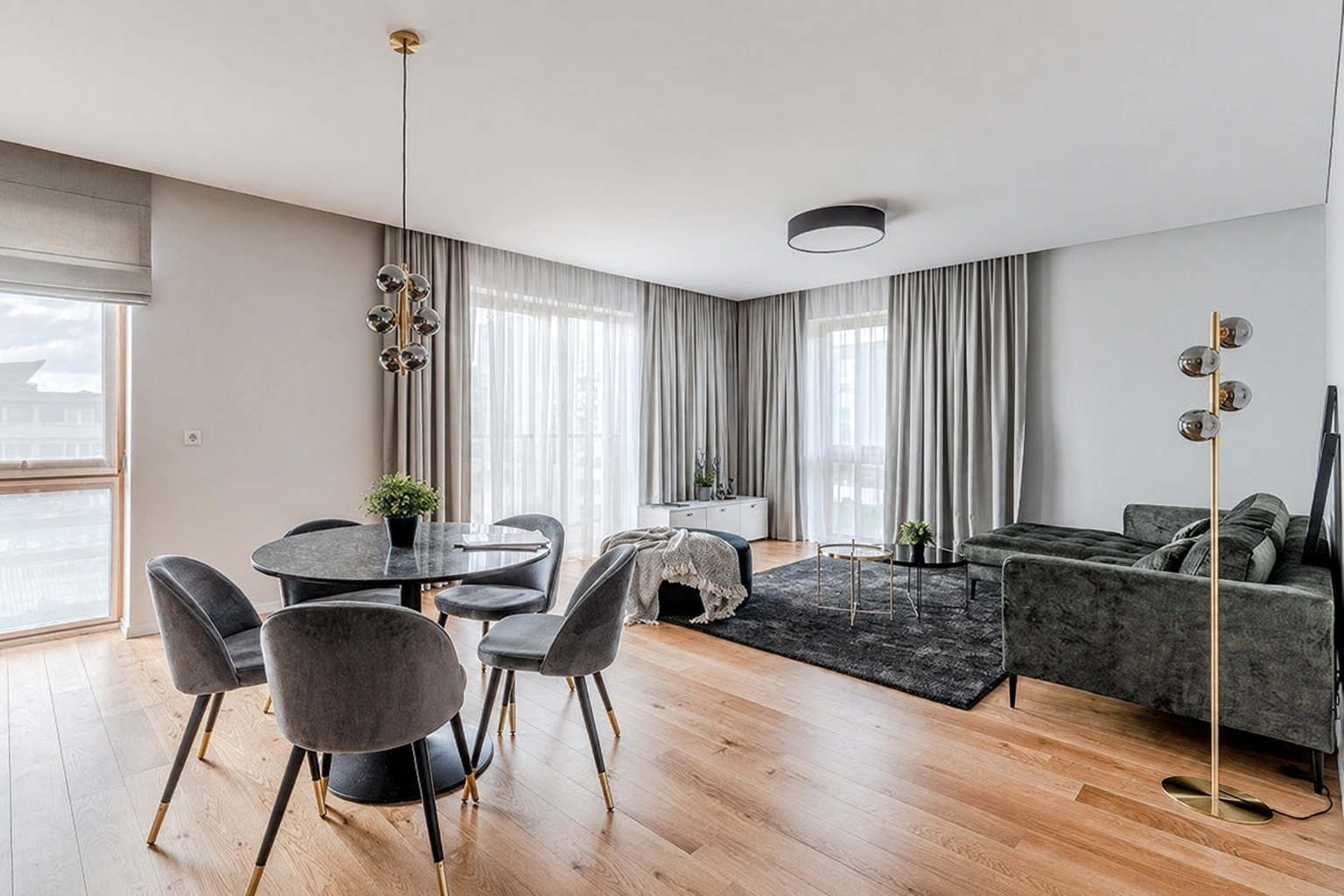 Im Wohnzimmer sind ein runder Esstisch, ein Sofa und große Fenster mit Vorhängen zu sehen.