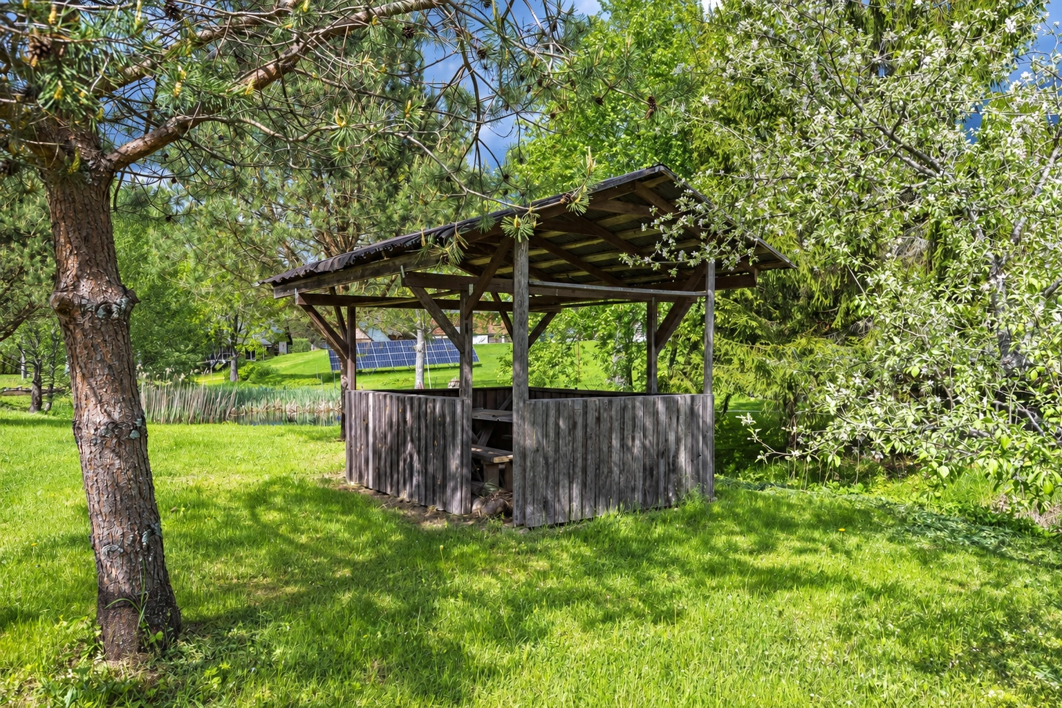 A gazebo with a roof and low walls stands on a lawn among trees.