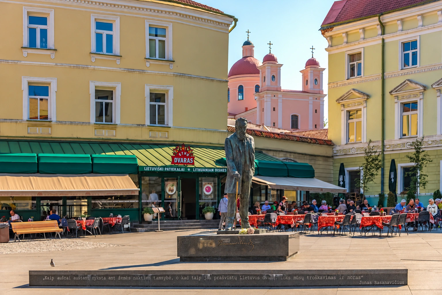 A sculpture of a person stands in the square, and outdoor café tables and yellow buildings can be seen around it.
