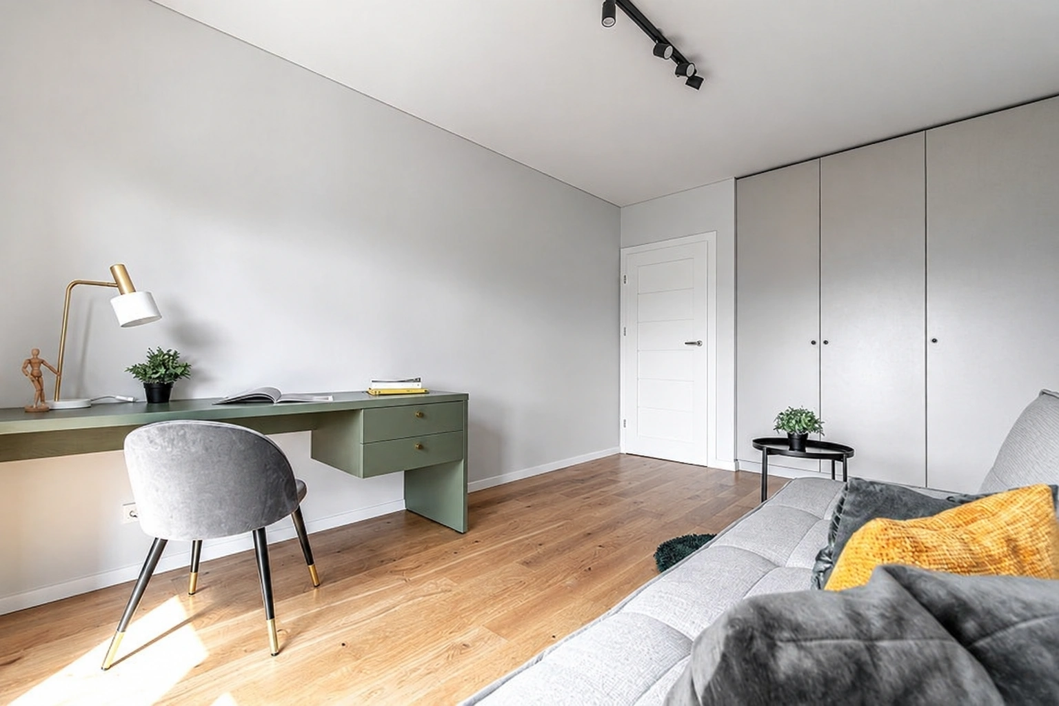 Ein Zimmer mit einem Schreibtisch und einem Stuhl, einem grauen Sofa sowie hohen Schränken.