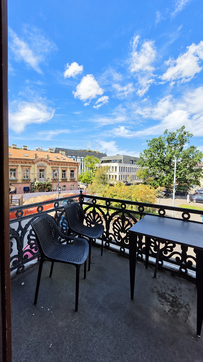 There are two chairs and a table on the balcony; trees and buildings can be seen beyond the openwork railings.