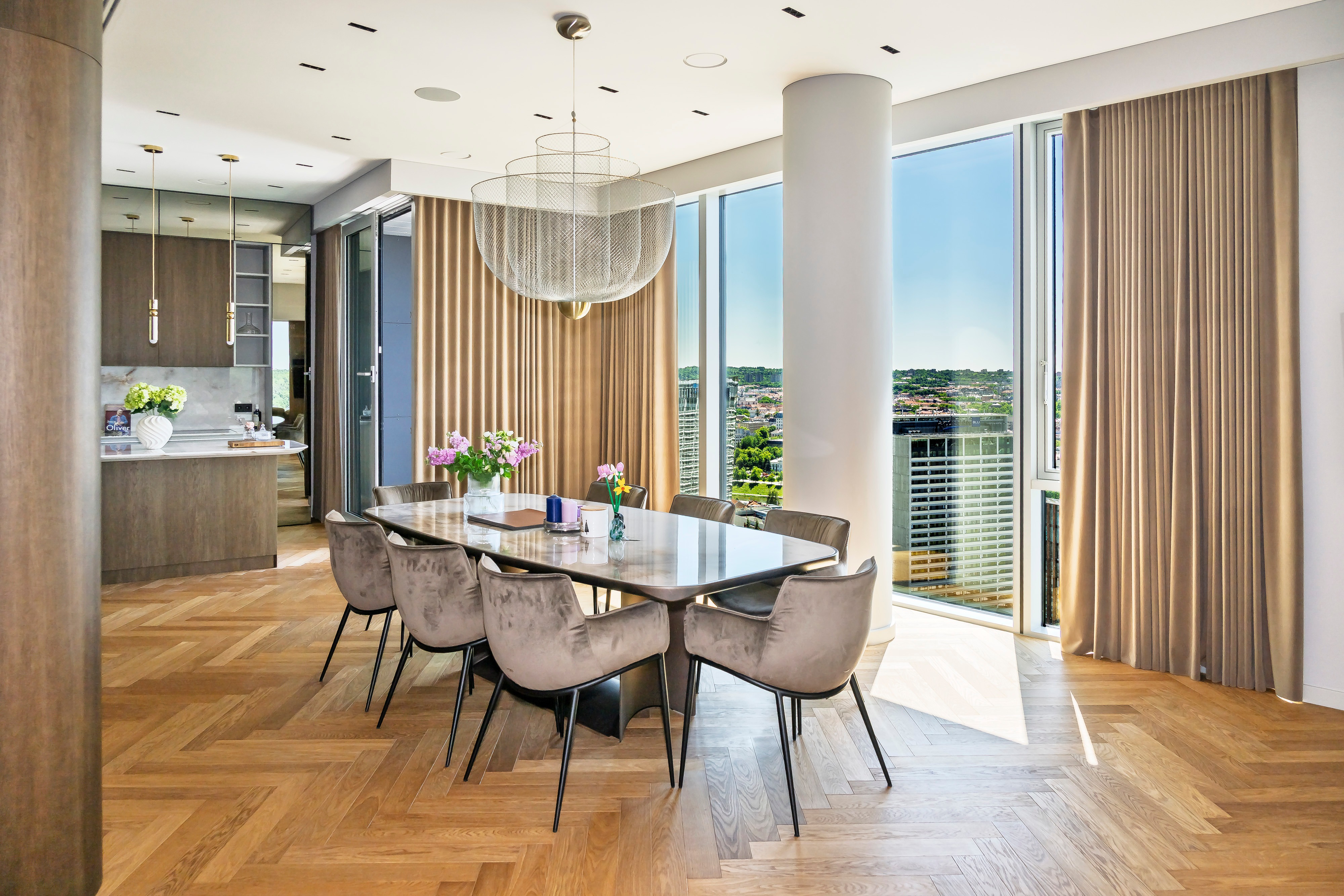 A dining table and upholstered chairs stand beside large windows with curtains.