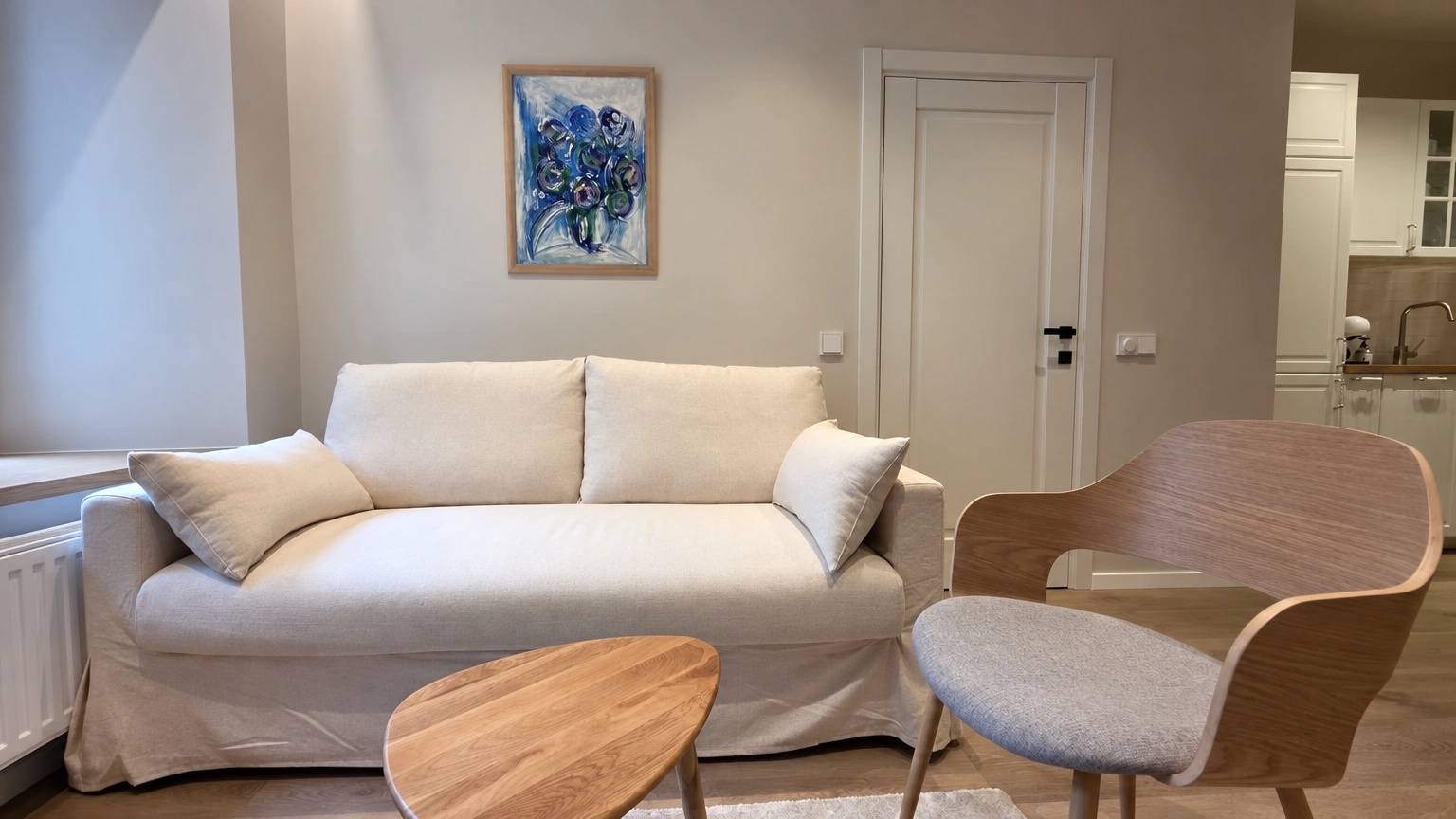 Svetainėje stovi šviesi sofa, krėslas ir kavos staliukas, ant sienos kabo paveikslas.