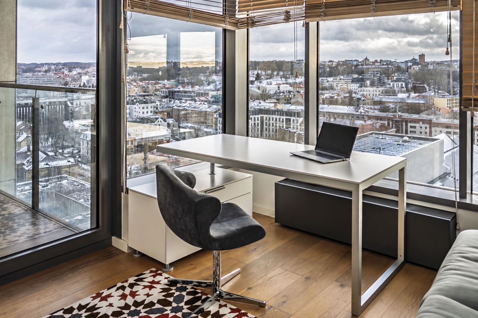By the large windows are a desk with a laptop and a black chair.