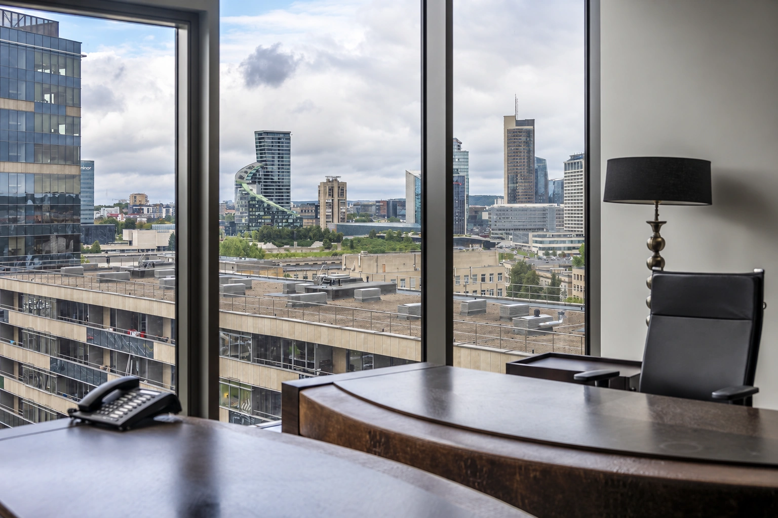 A desk and a black chair stand by the large windows, and city buildings are visible outside the windows.