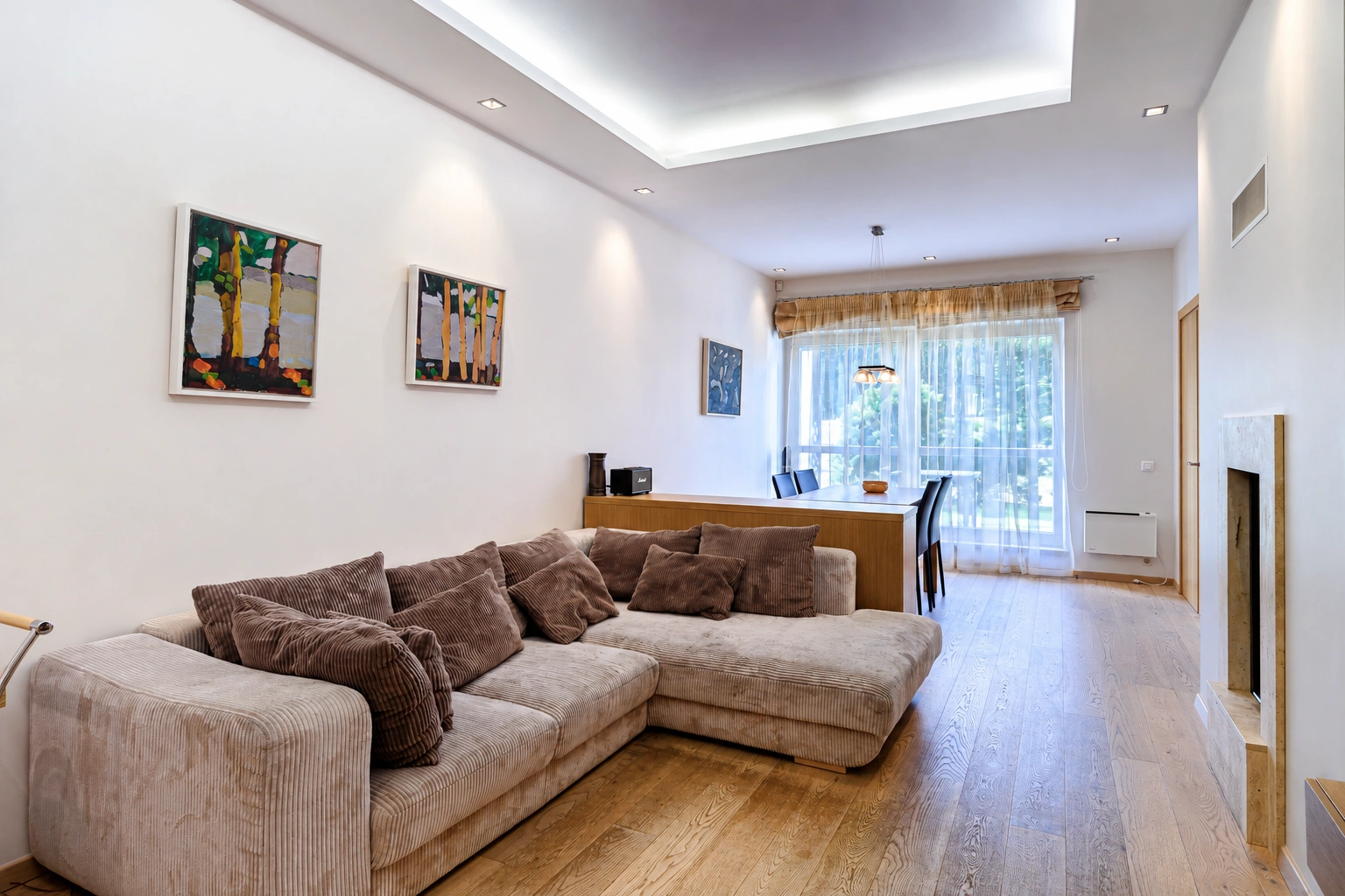 Svetainėje stovi kampinė sofa su pagalvėlėmis, ant sienos kabo paveikslai.
