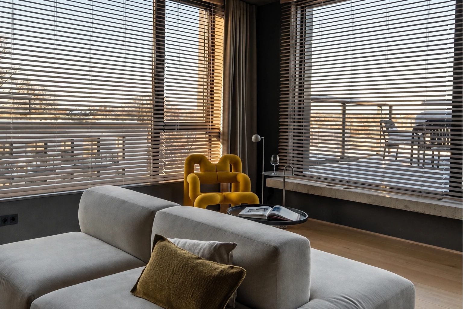 Svetainėje stovi šviesi sofa ir geltonas fotelis, o didelius langus dengia horizontalios žaliuzės.