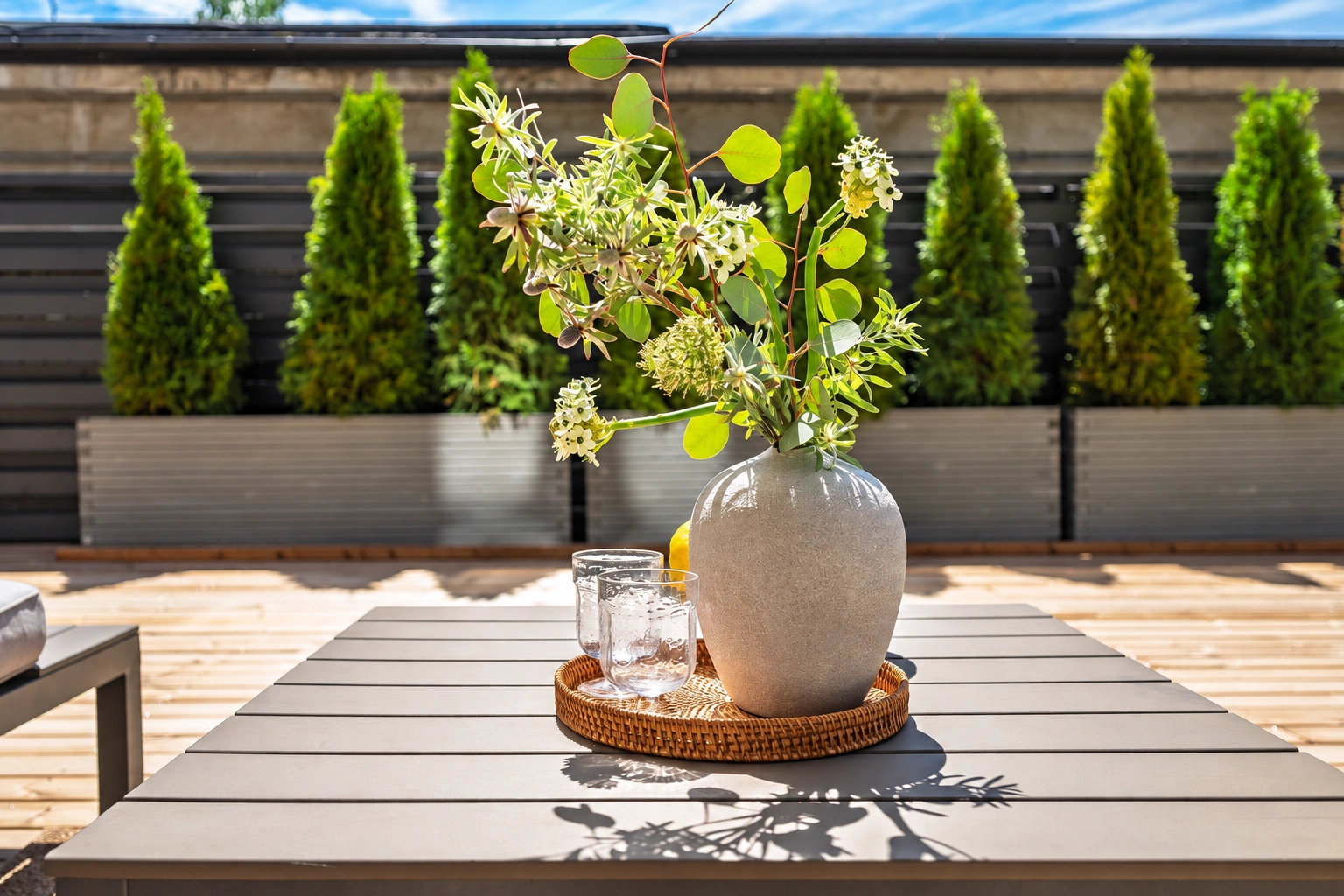 Auf dem Terrassentisch steht eine Vase mit blühenden Zweigen; im Hintergrund sind in Töpfen wachsende Thujen zu sehen.
