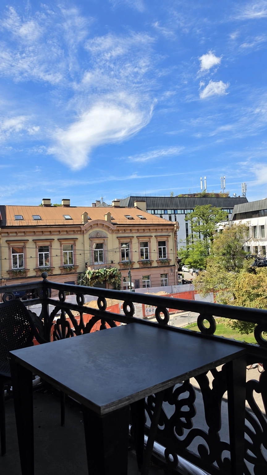 There is a table on the balcony, and city buildings and trees can be seen beyond the decorative railing.