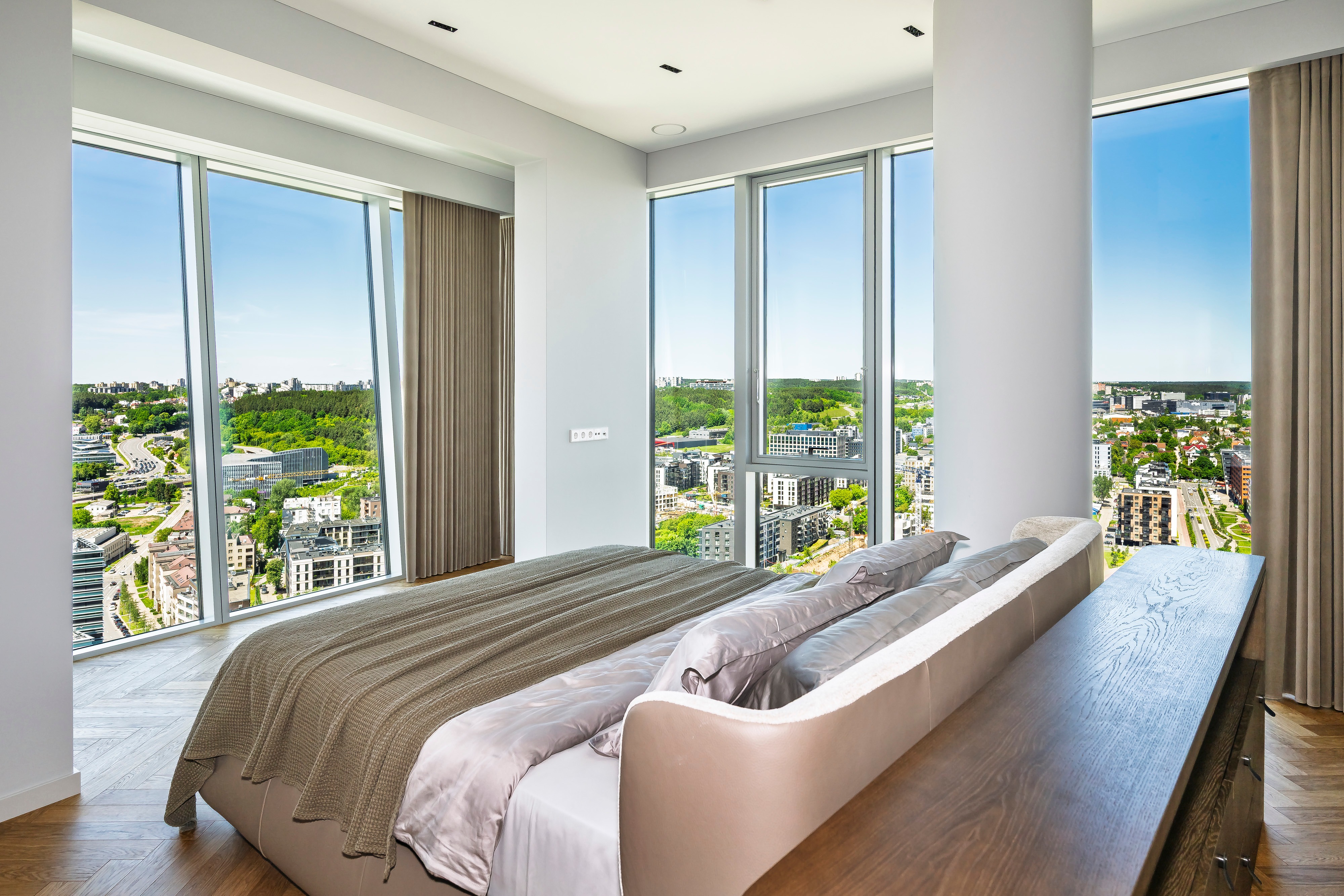 Bedroom with a bed and large windows through which city buildings and trees can be seen.