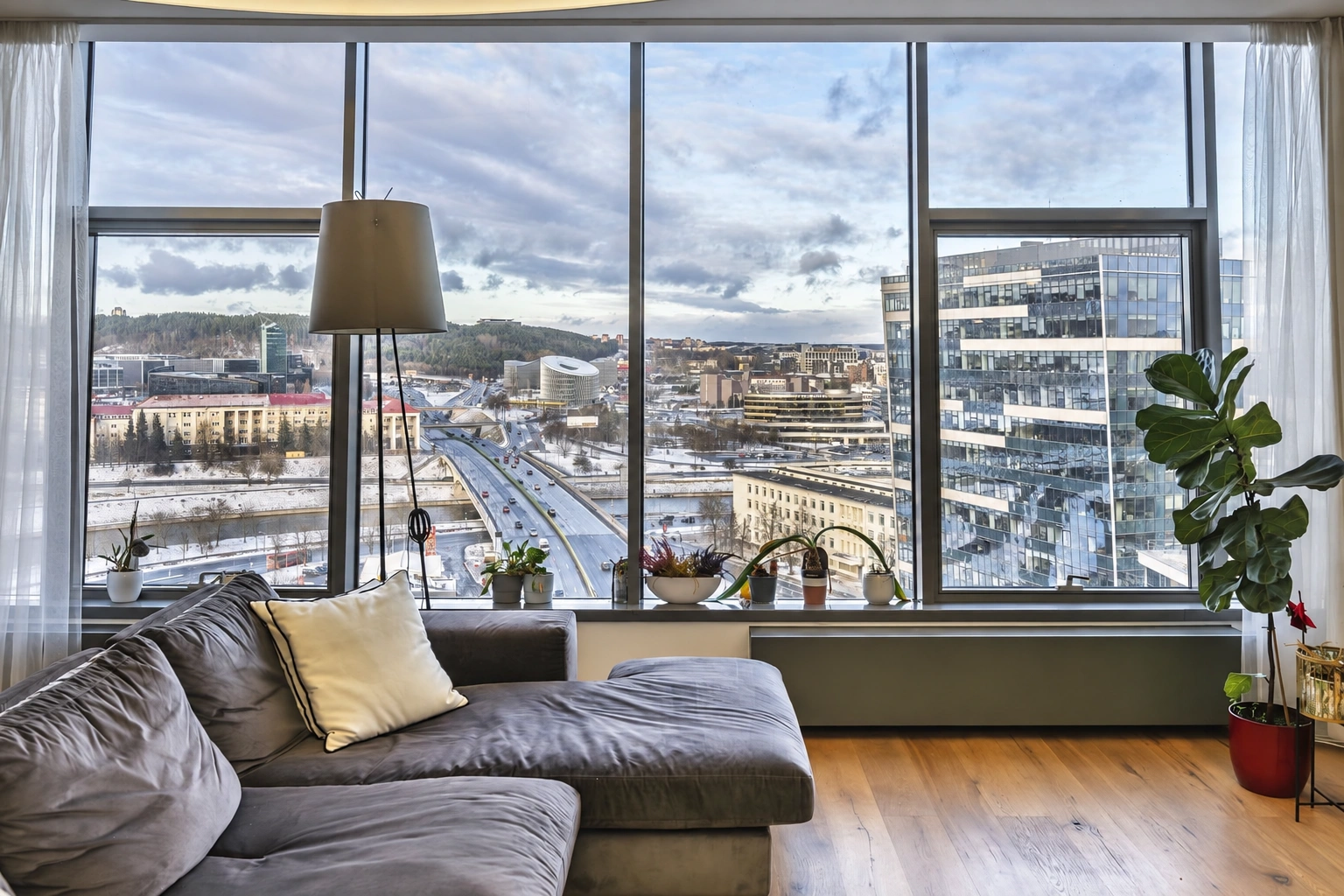 Wohnzimmer mit einem grauen Sofa und großen Fenstern, durch die Gebäude der Stadt zu sehen sind.
