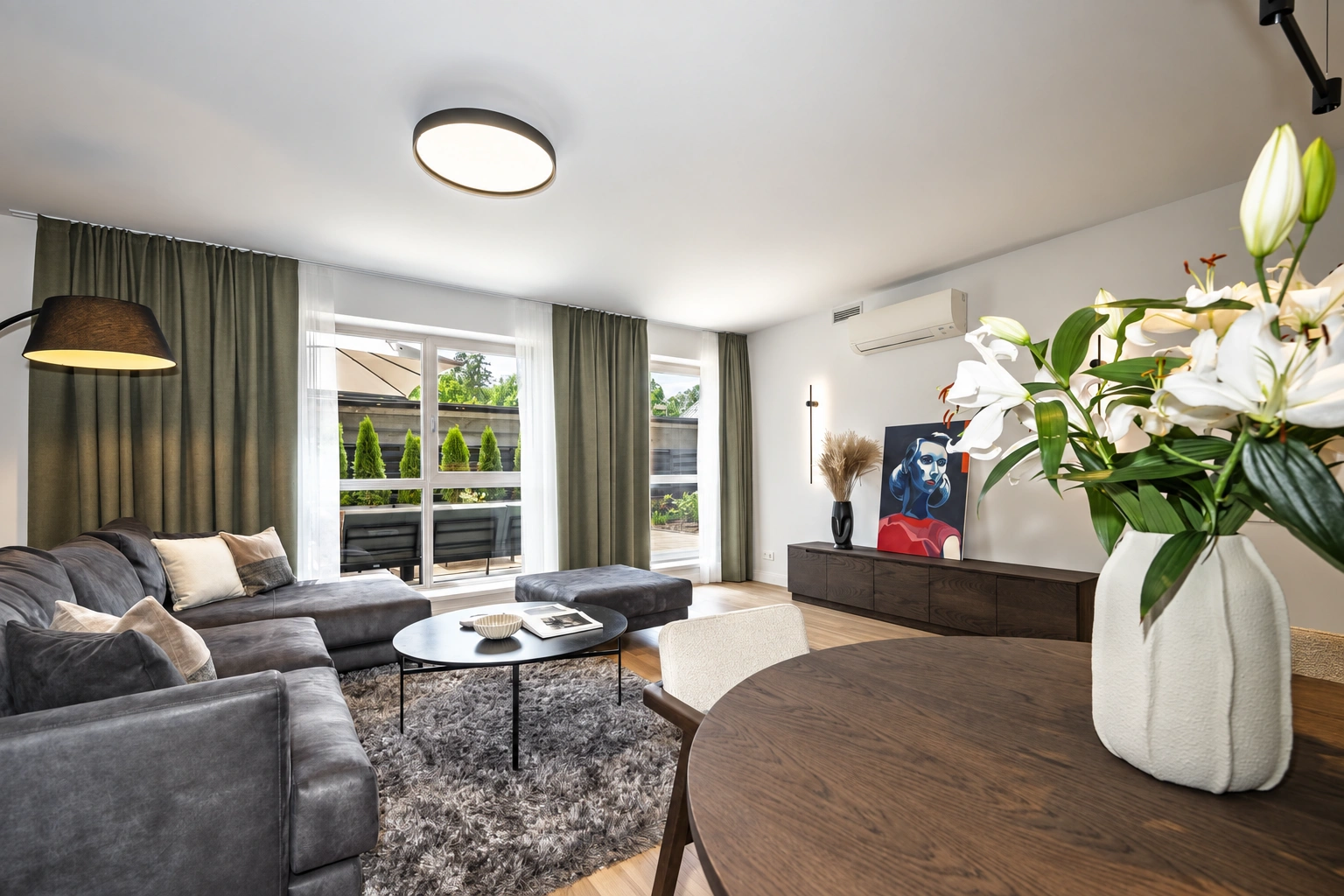 Im Wohnzimmer stehen ein graues Sofa, ein Couchtisch und ein Esstisch mit einer Blumenvase.