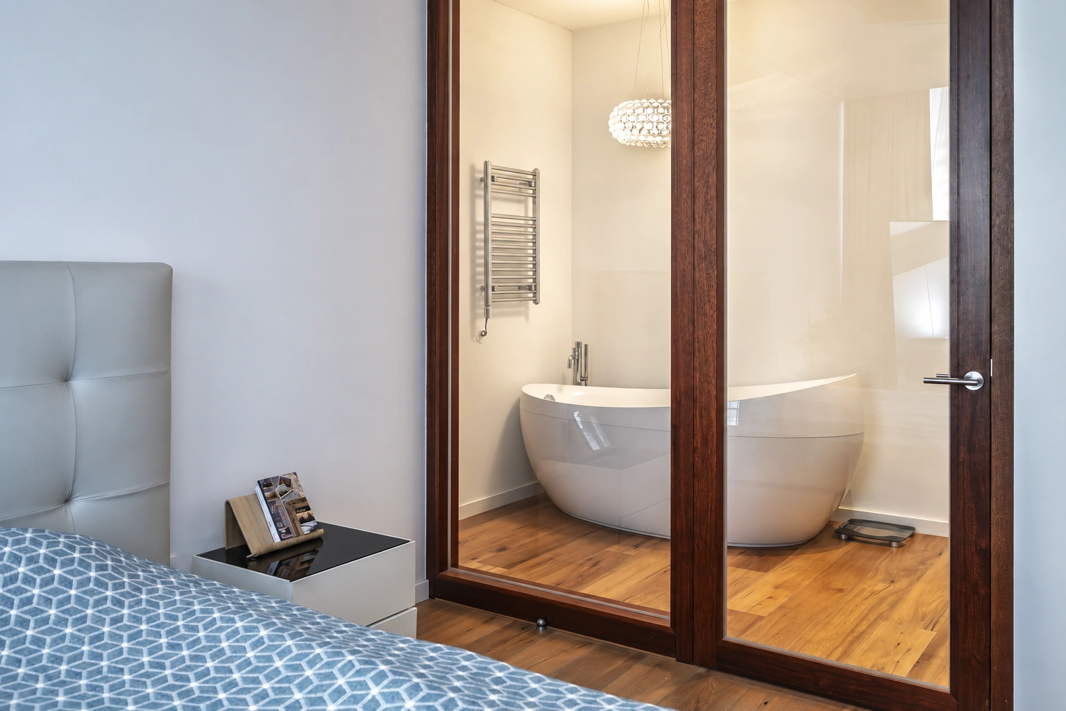 There is a bed in the bedroom, and a freestanding white bathtub is visible behind the glass doors.