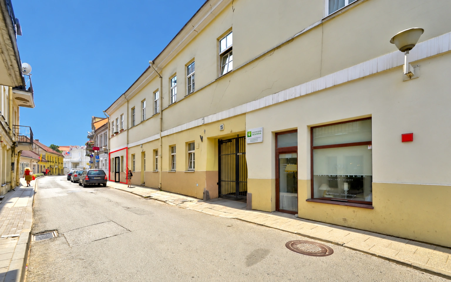 An einer schmalen Straße steht ein gelbliches Gebäude mit großen Fenstern und einer Eingangstür.
