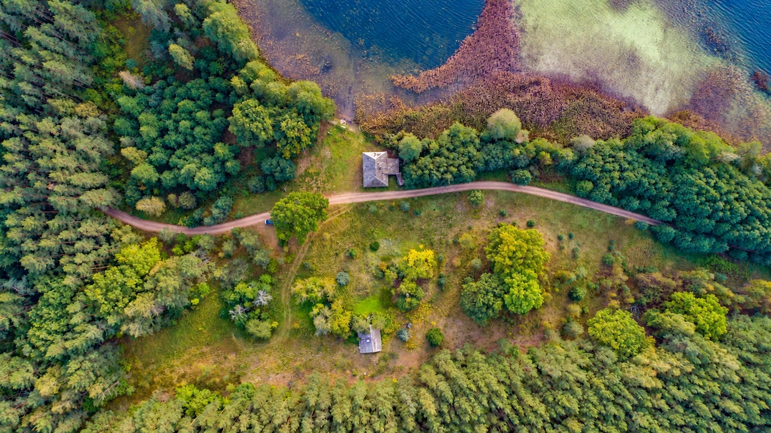 Iš viršaus matoma medžiais apsupta pieva su keliuku ir vandens telkiniu šalia.