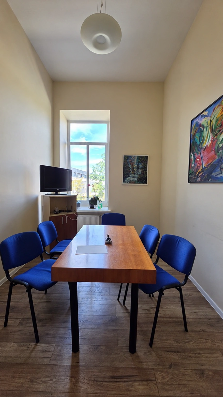 A rectangular table with blue chairs stands in a narrow room, and there is a window at the end.