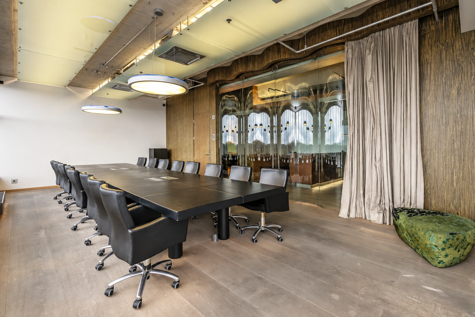 There is a long table with black chairs in the room, and light-colored curtains hang by the glass partition.