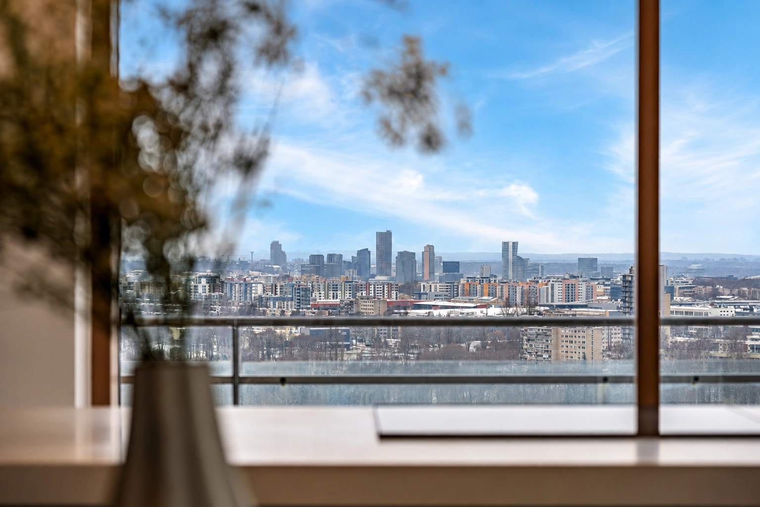 Durch die großen Fenster ist ein Stadtpanorama zu sehen, und im Vordergrund steht eine Vase mit Zweigen.