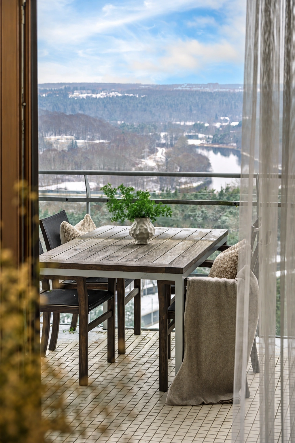 Auf dem Balkon stehen ein Tisch mit einer Topfpflanze und Stühle; hinter dem Geländer sind bewaldete Hänge zu sehen.