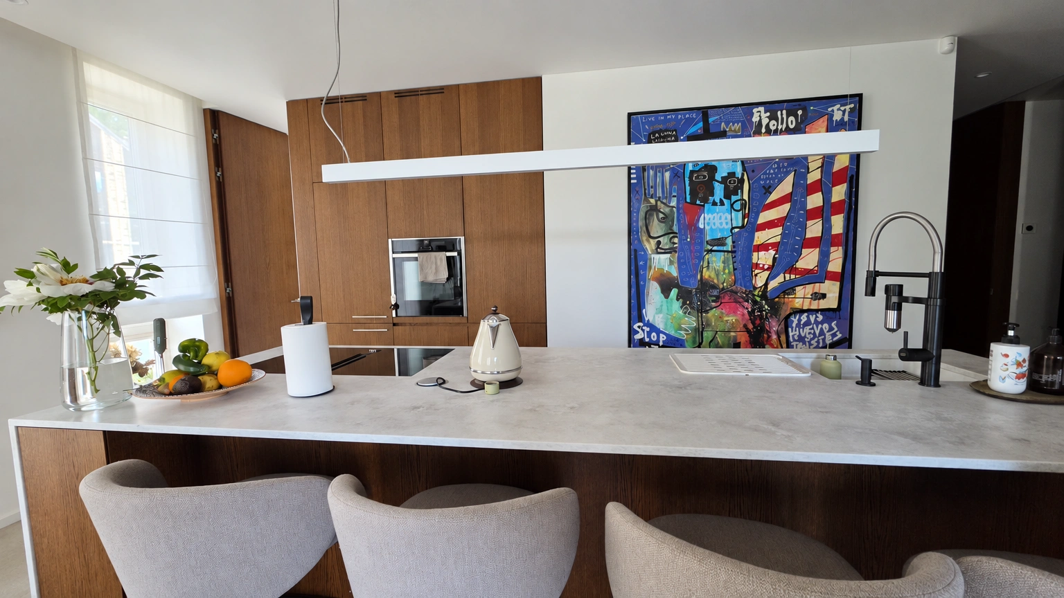 Kitchen with an island, tall chairs and a large, colourful painting by the wall.
