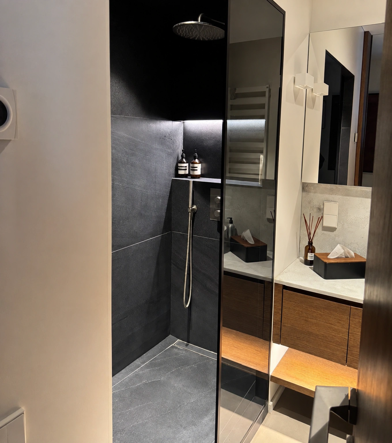 In the bathroom, a shower with a glass partition, a washbasin, and a mirror above it can be seen.