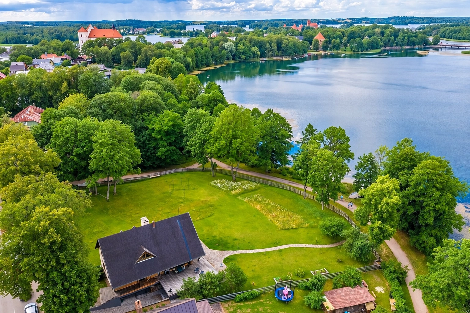 Iš viršaus matyti ežero pakrantė su medžiais ir namu tamsiu stogu.