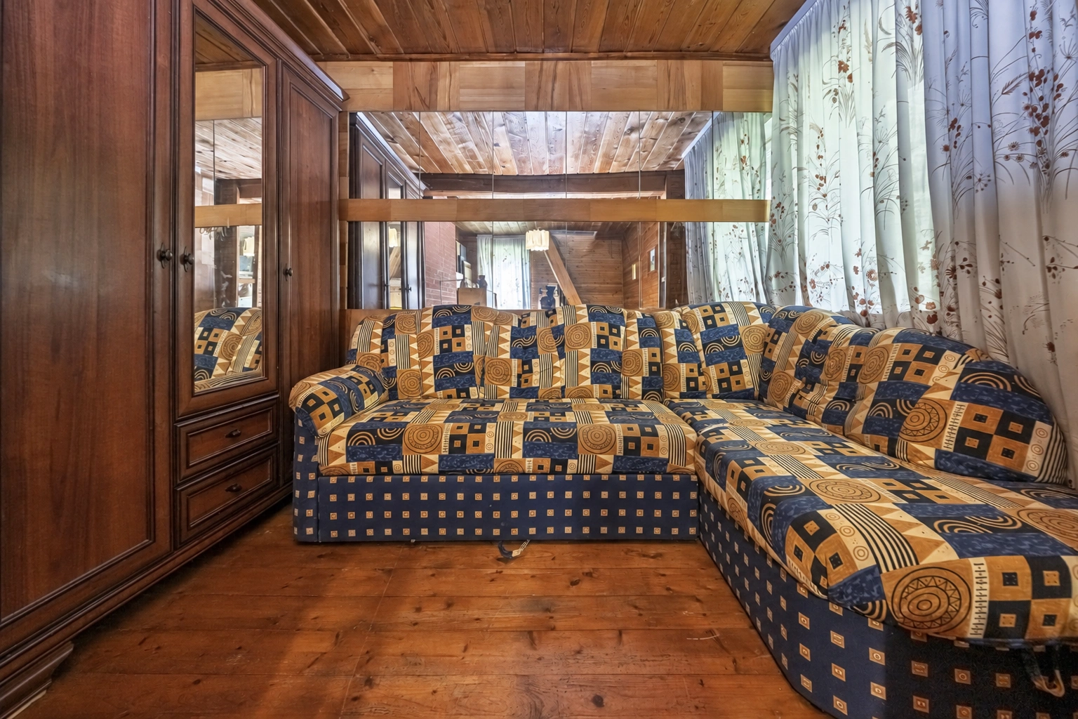 A room with a patterned corner sofa and windows covered with light-colored curtains.