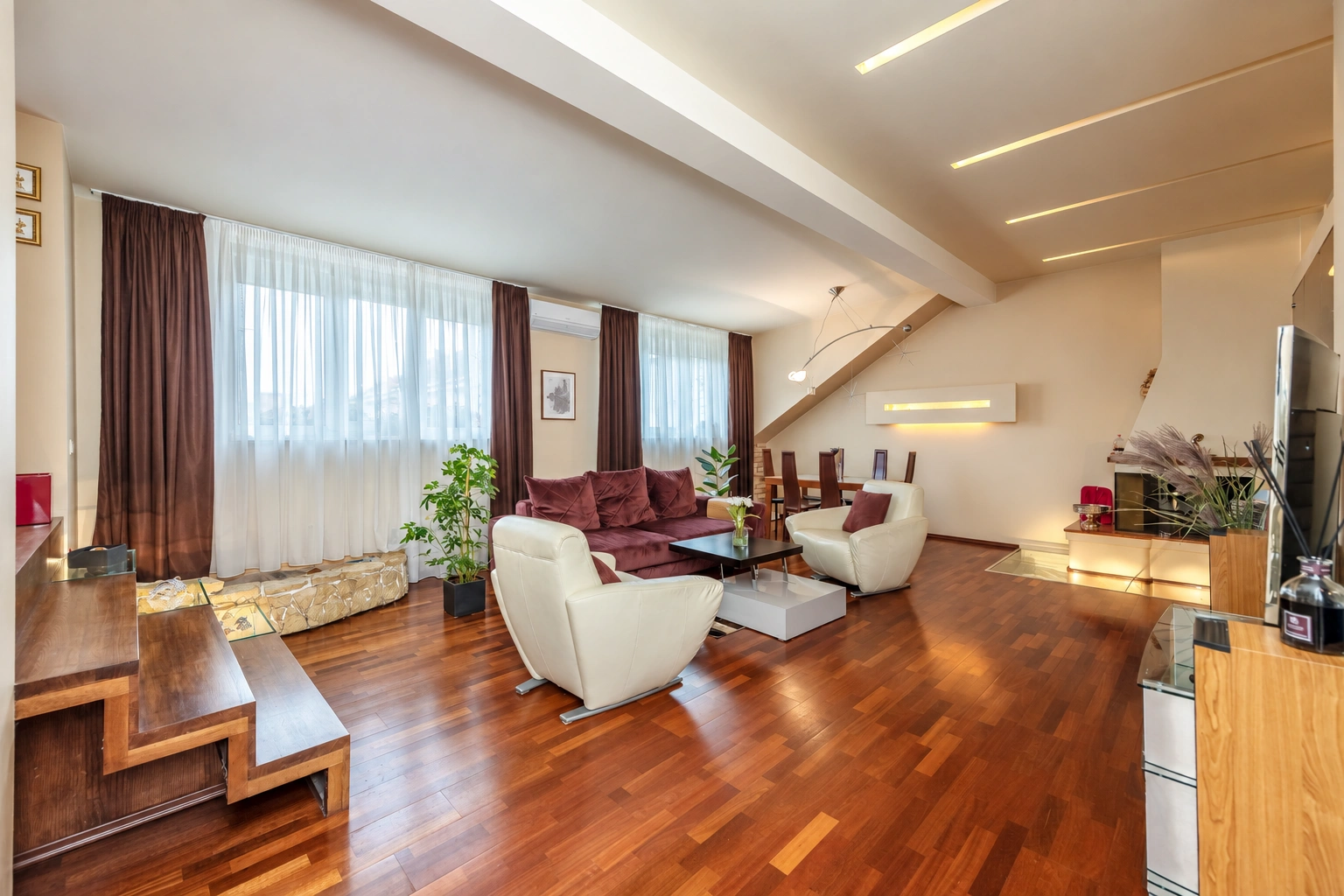 Svetainė su sofa ir foteliais, dideliais langais bei laiptais į viršutinį aukštą.