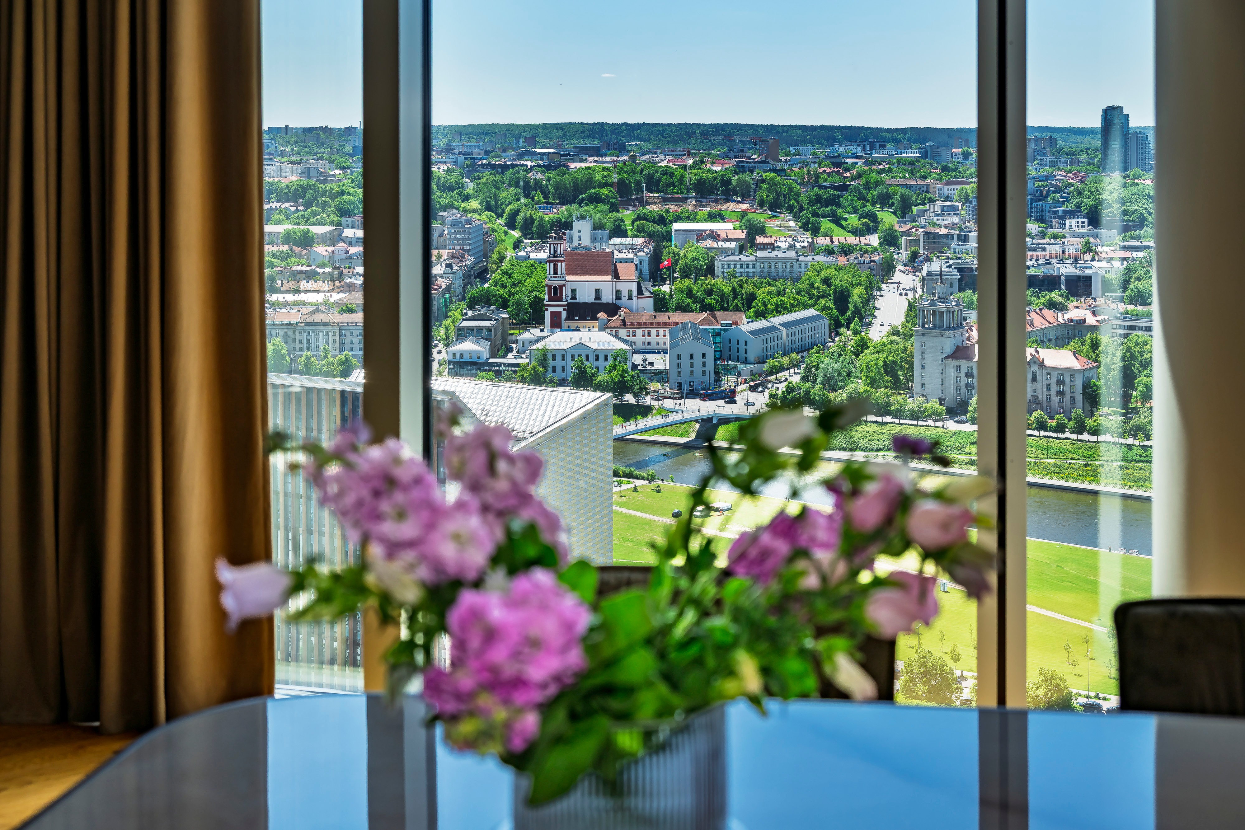 A vase of flowers stands on the table beside large windows, and city buildings can be seen outside the windows.