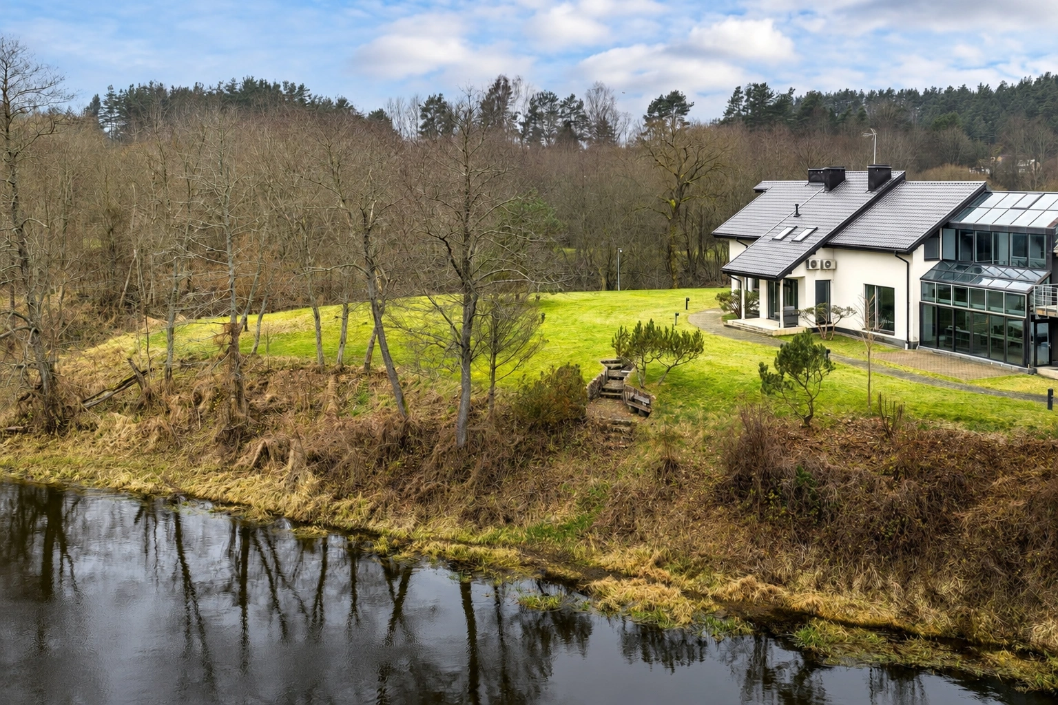 In der Nähe eines Gewässers steht ein Haus mit großen Fenstern; um das Haus herum sind eine Rasenfläche und Bäume zu sehen.