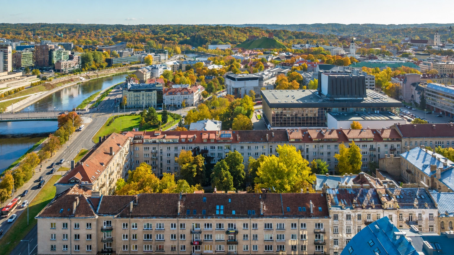 Miesto panorama iš viršaus su gyvenamaisiais pastatais, upe ir rudeniškai nusidažiusiais medžiais.