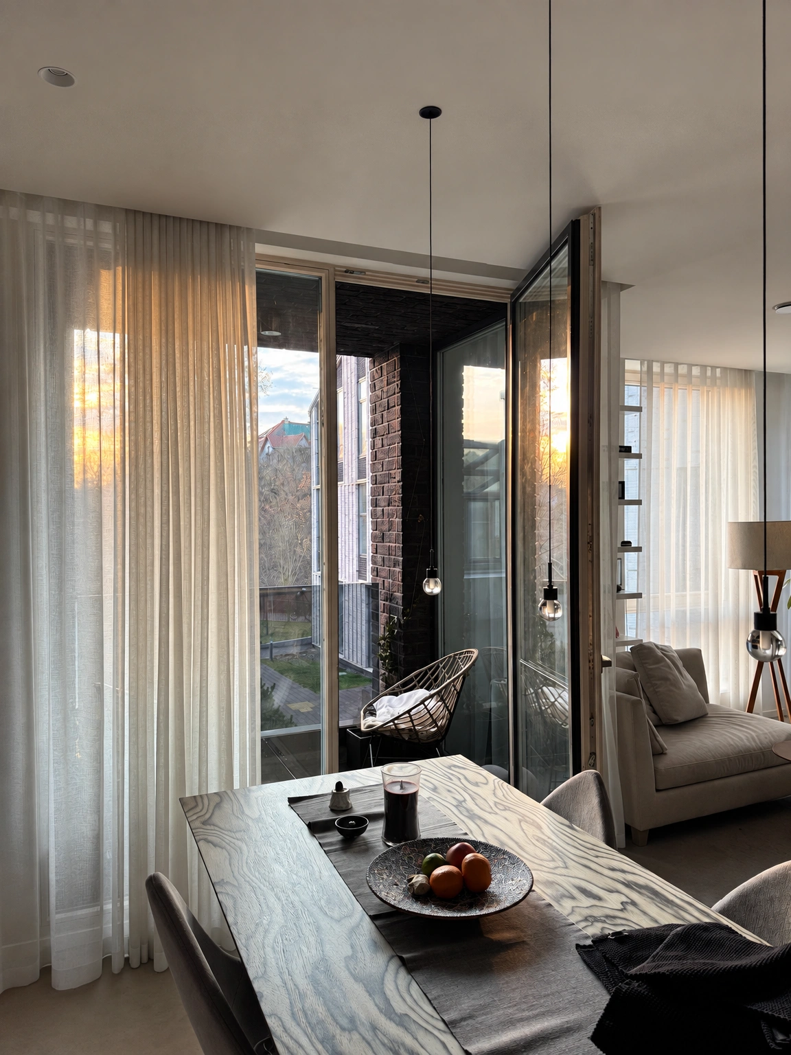 A dining table with chairs stands by the windows with light-colored curtains; light fixtures hang above it.