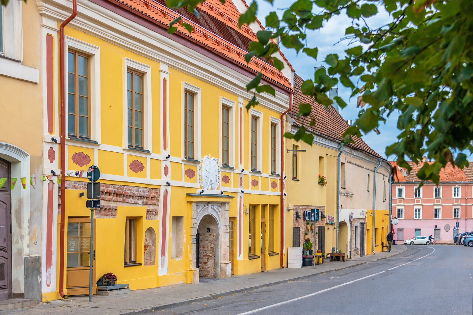 Palei gatvę stovi geltoni pastatai su raudonais stogais, viršuje matyti medžio šakos.