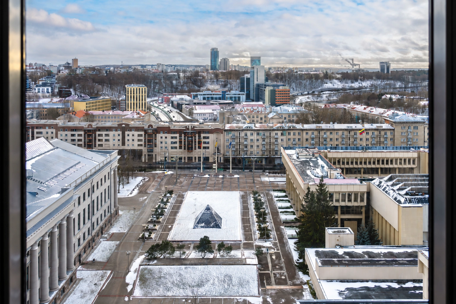 Through the window, a snow-covered courtyard with paths and a panoramic view of the city can be seen.