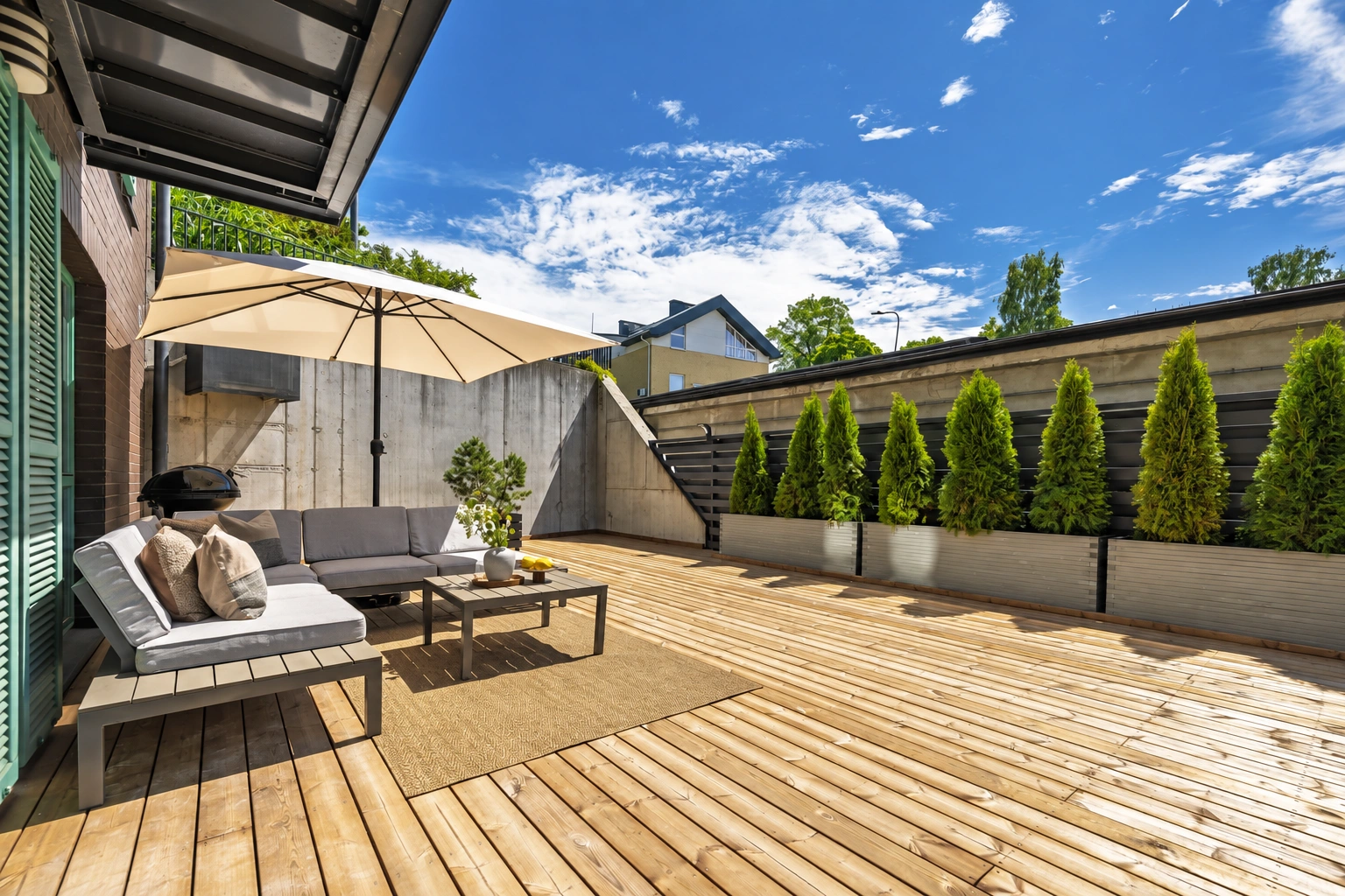 Eine Terrasse mit Gartenmöbeln unter einem Sonnenschirm und einer Reihe von Nadelbäumen entlang des Zauns.