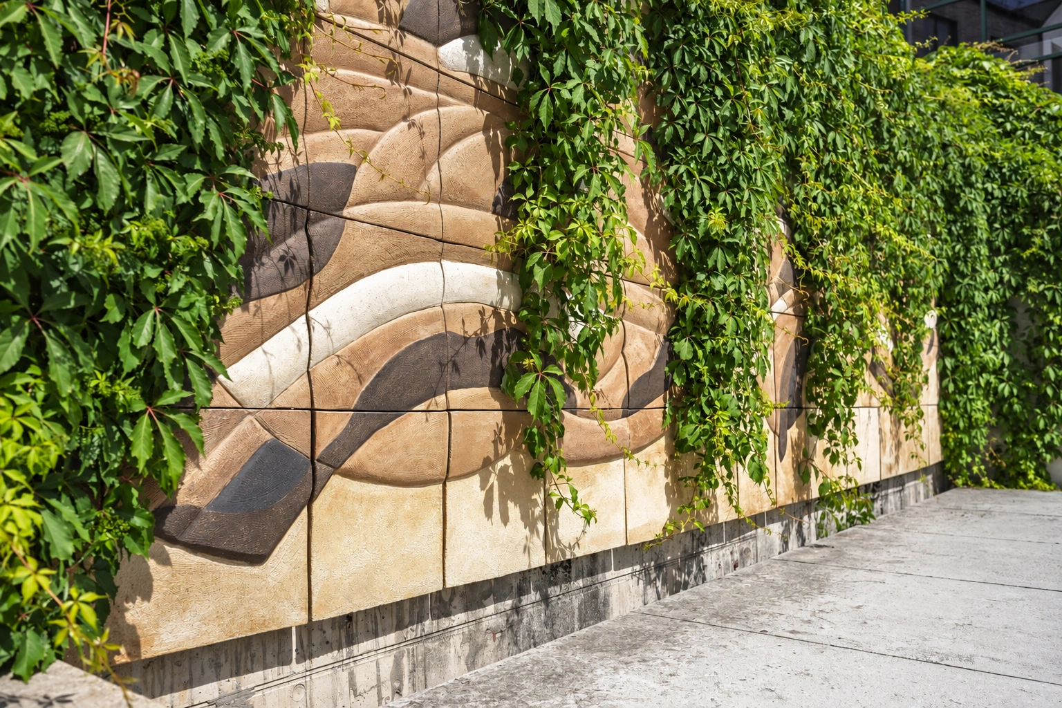 Eine Wand mit einem wellenförmigen Muster ist von Kletterpflanzen überwachsen, und entlang der Wand verläuft ein Gehweg.