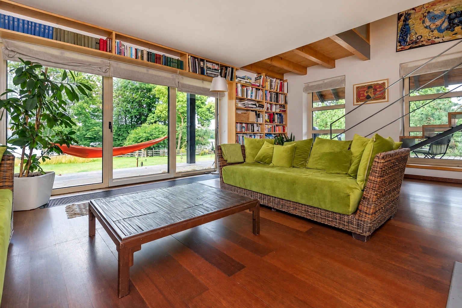 Svetainė su žalia sofa ir dideliais langais, pro kuriuos matyti žaluma.