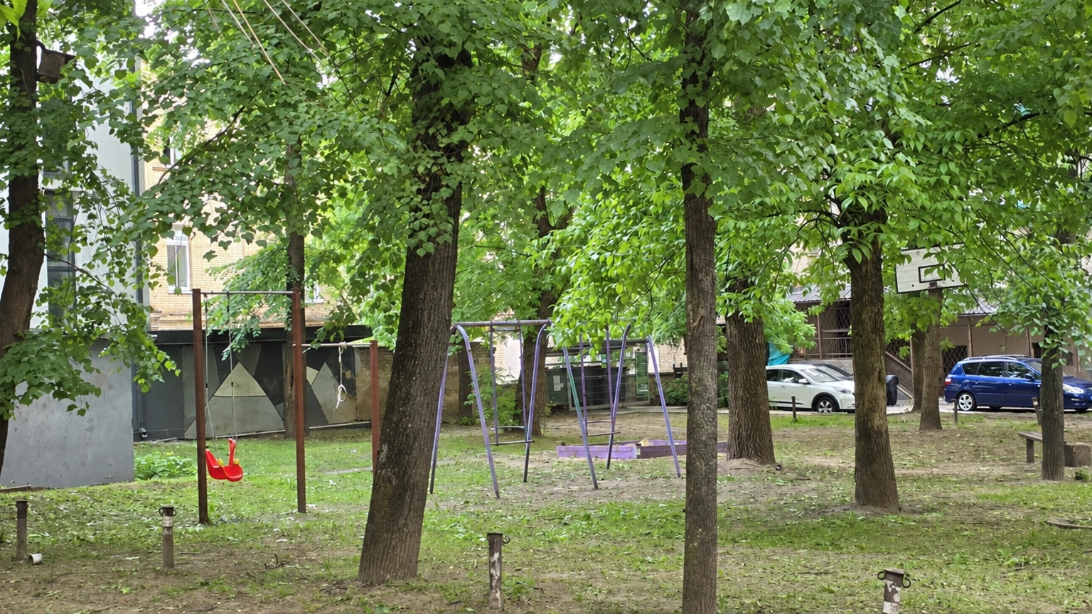 Kieme tarp medžių įrengtos sūpynės, o fone matyti pastatai.