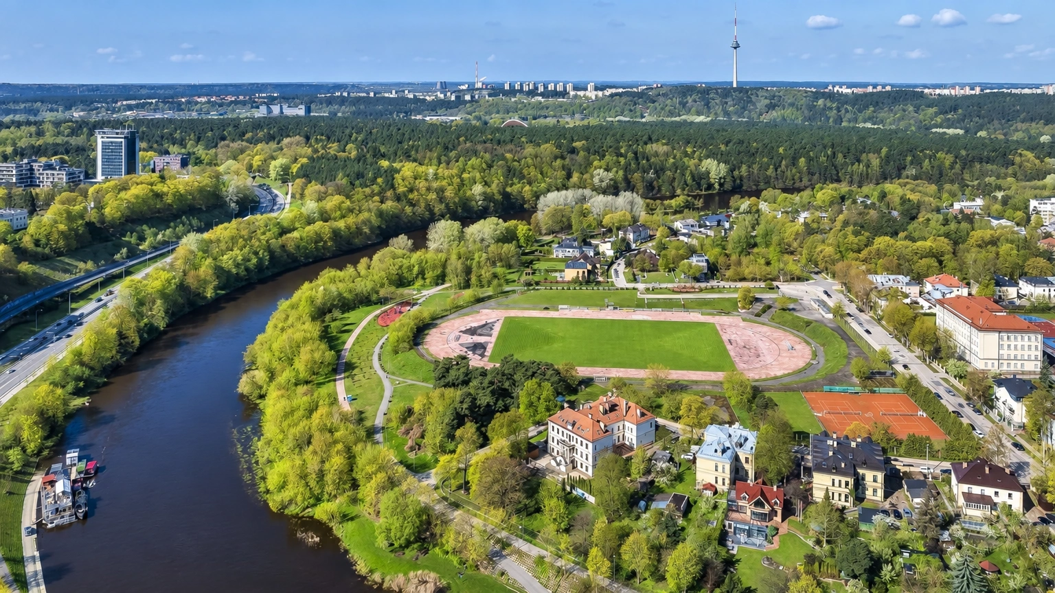 From above, a bend in the river, a stadium with a running track, and buildings among the trees are visible.