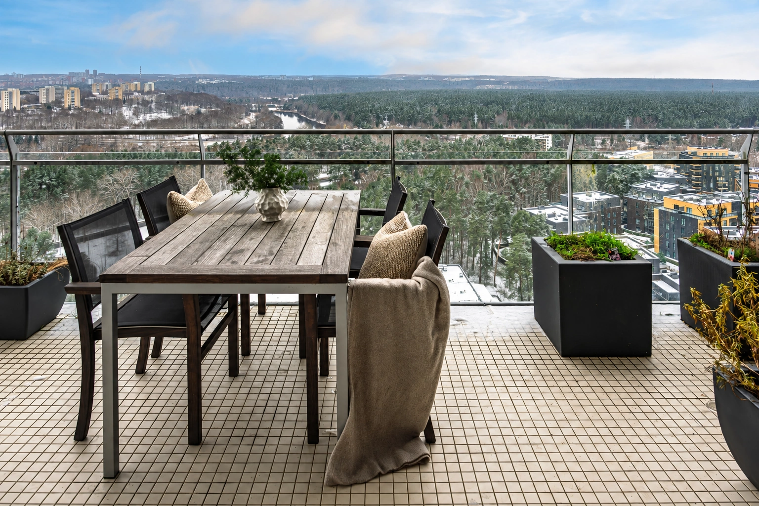 Eine Terrasse mit einem Tisch, Stühlen und Pflanzentöpfen; hinter dem Geländer sind Bäume zu sehen.