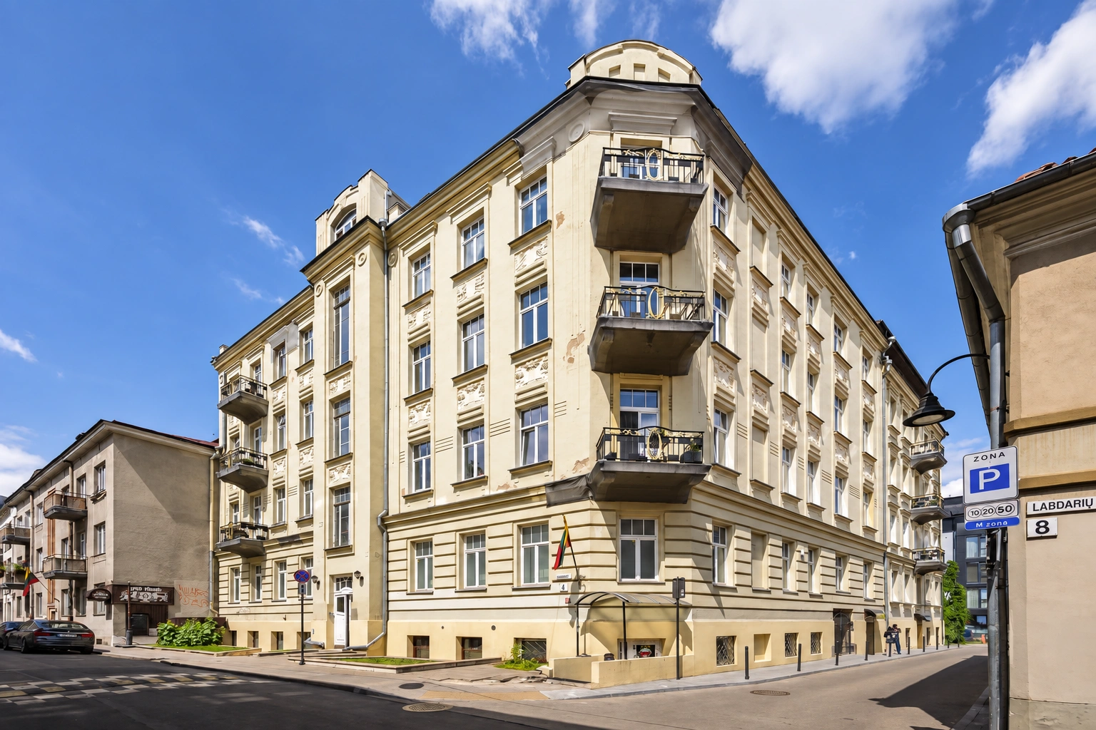 Gatvių sankirtoje stovi daugiaaukštis pastatas su balkonais ir eilėmis išdėstytais langais.