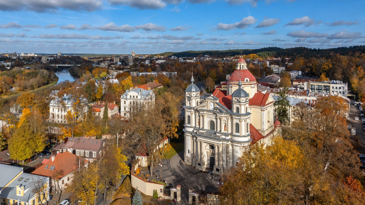 Bažnyčią su bokštais ir raudonu stogu supa rudeniniai medžiai ir miesto pastatai.