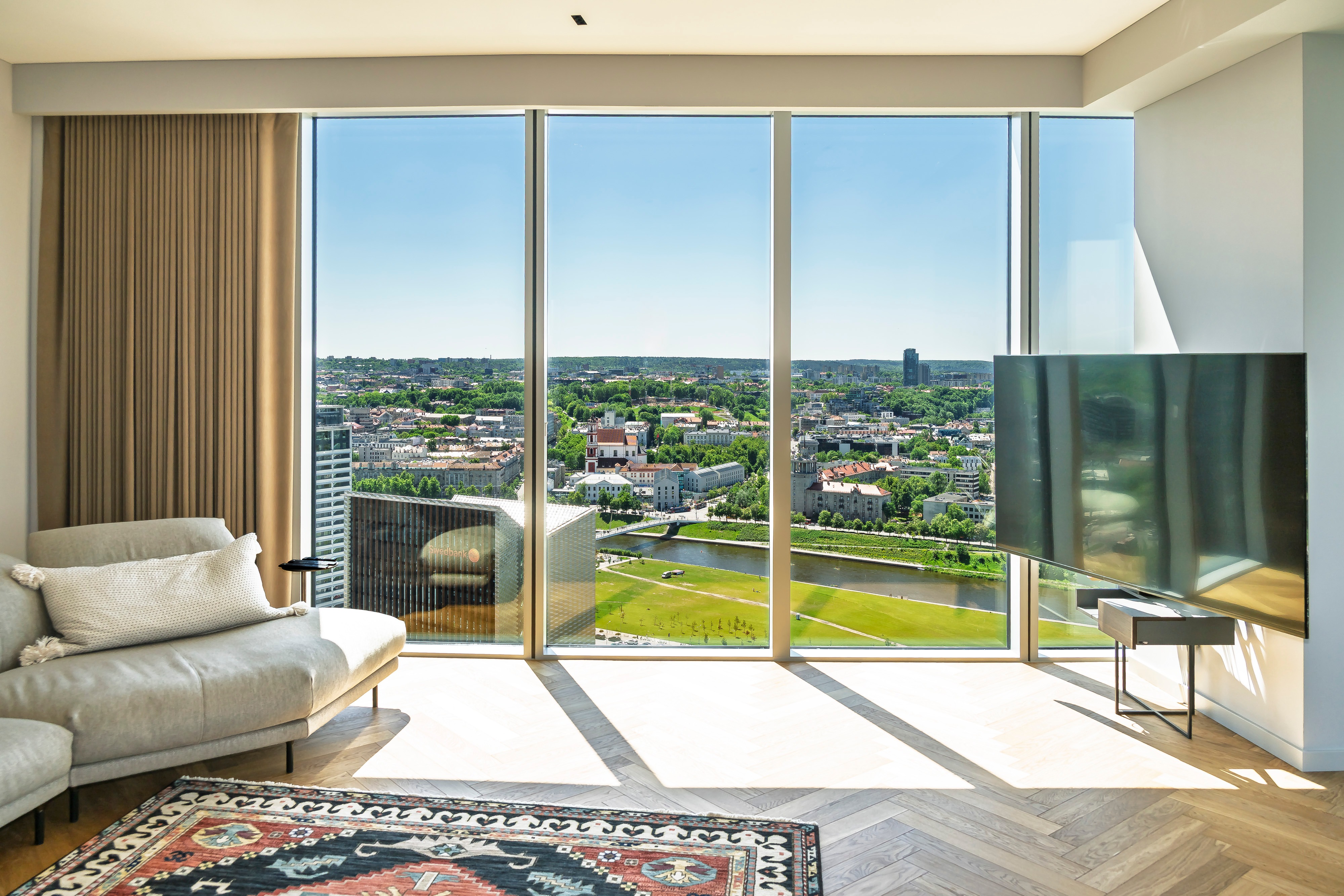 Living room with a sofa and large windows through which city buildings and green spaces can be seen.