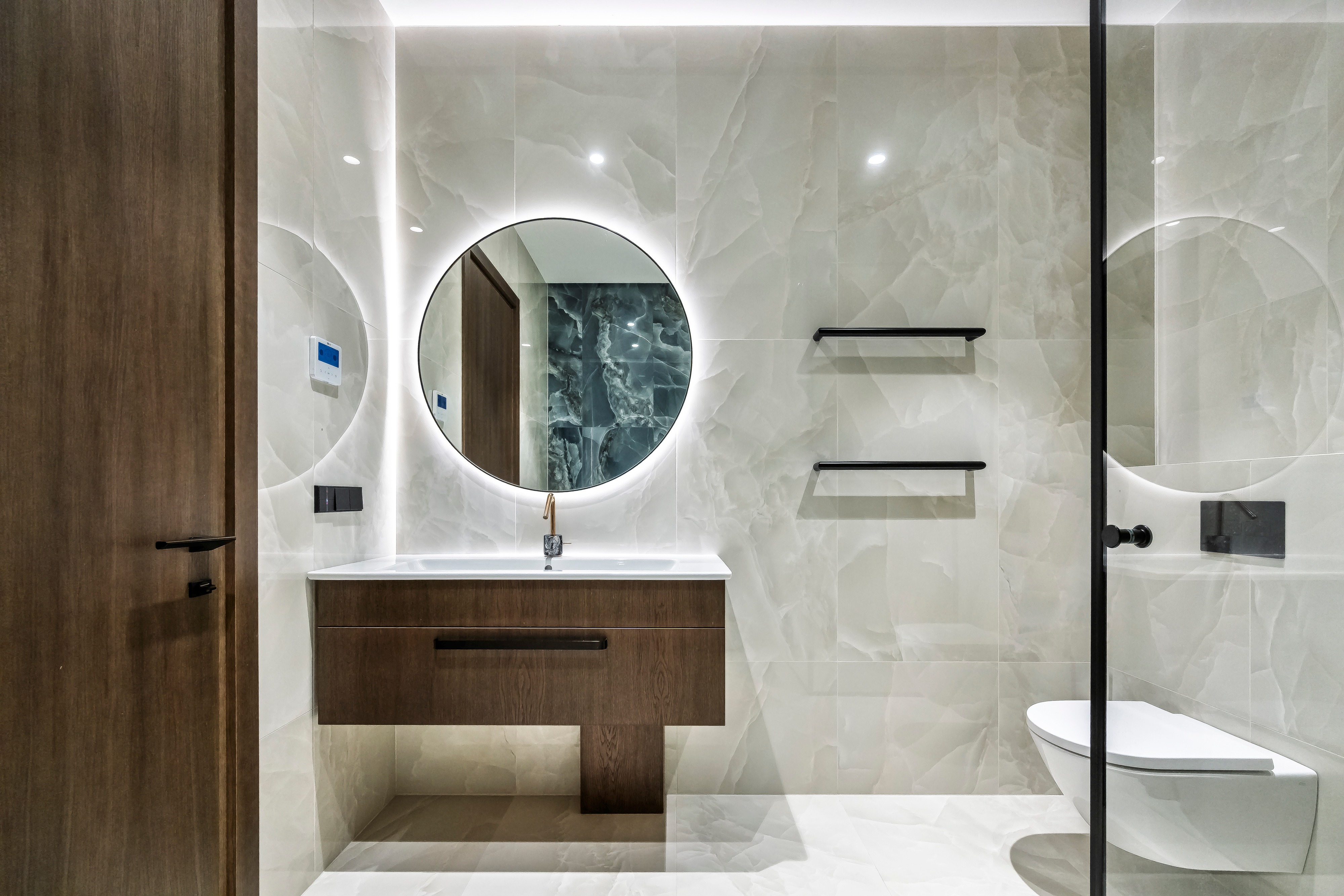Bathroom with a washbasin, a round illuminated mirror, wall shelves and a glass partition.