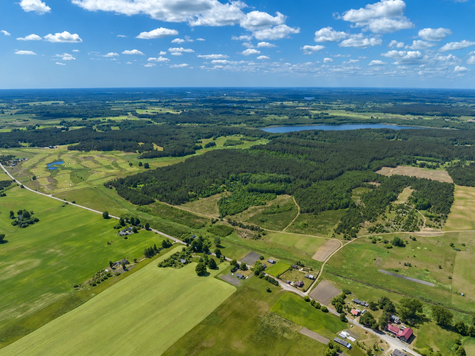 Aus der Luft sind Felder und Wälder zu sehen, zwischen denen sich Straßen erstrecken; in der Ferne ist ein See sichtbar.