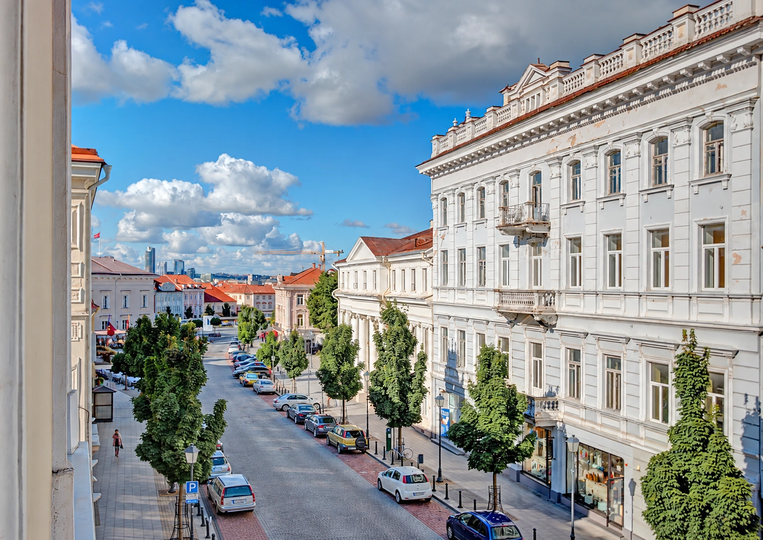 Palei šviesų daugiaaukštį pastatą driekiasi gatvė su medžiais ir stovinčiais automobiliais.