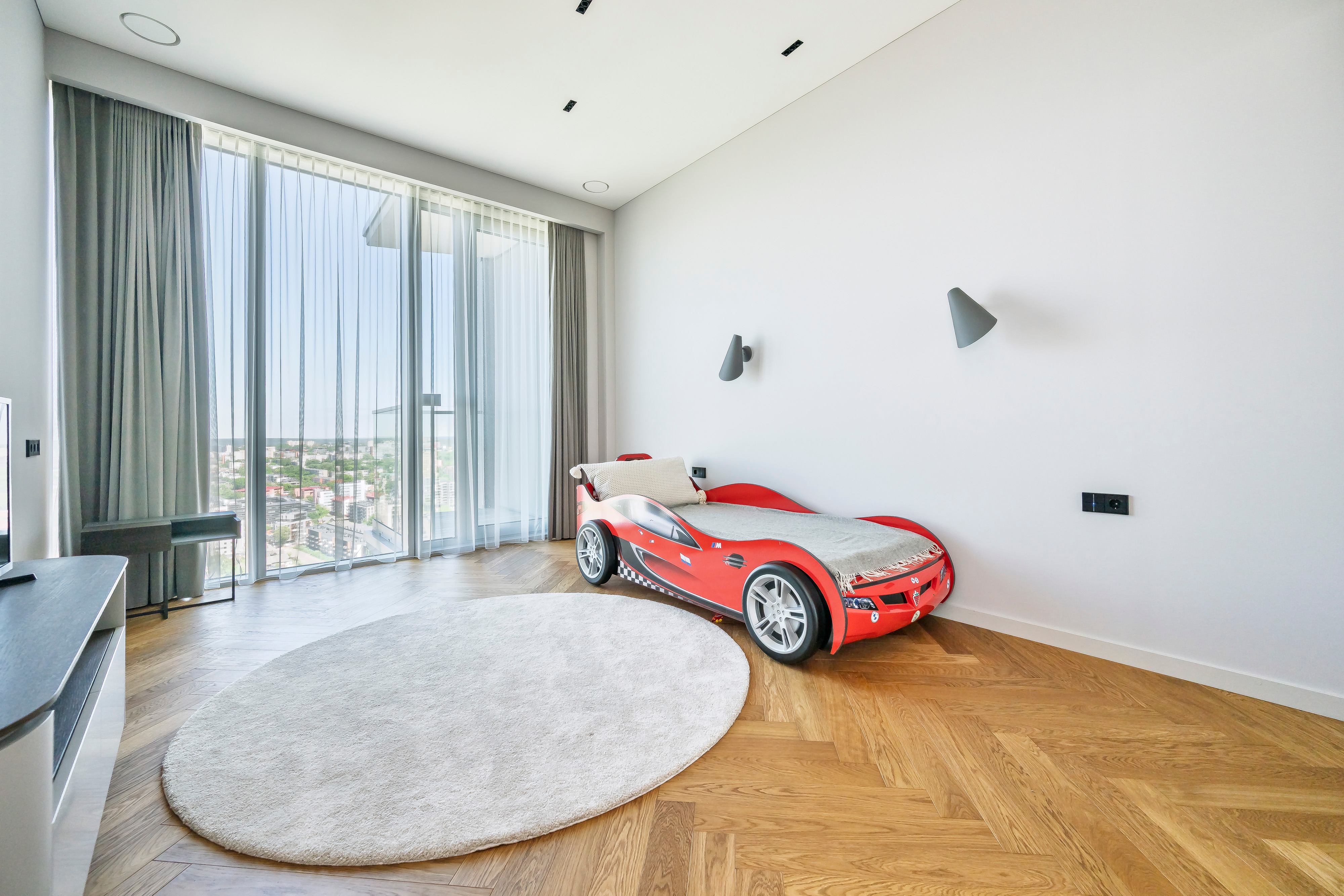 Room with a red car-shaped bed, a round rug and large floor-to-ceiling windows.